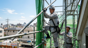 灼熱の7月の太陽の下、空調服を着用し、ヘルメットから汗を流しながらも、真剣な眼差しで鉄パイプを組み立てる足場鳶の職人。背景には、夏の強い日差しに照らされたメッシュシートと、青い空が広がる。職人の手元には、スポーツドリンクのボトルと、WBGT測定器が置かれ、暑さと戦いながらも安全を最優先している様子が伝わる実写画像。